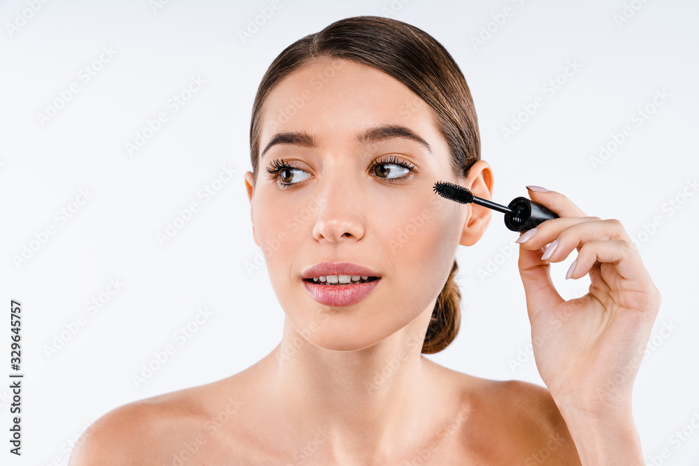 Beautiful young woman with makeup brush applying black mascara on eyelashes isolated on white background.