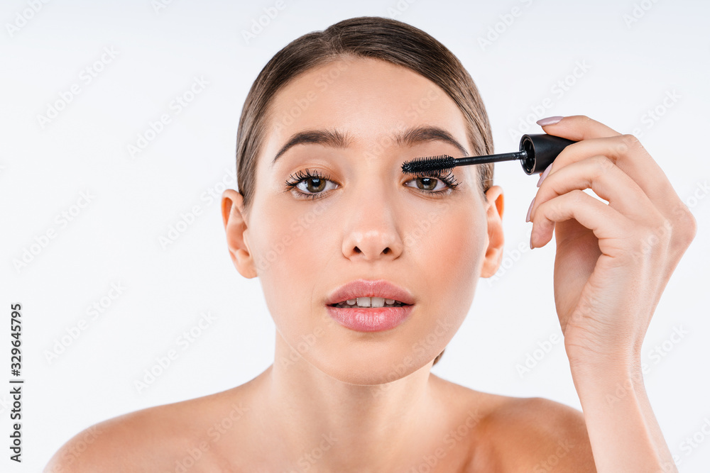 Portrait of young woman with makeup brush applying black mascara on eyelashes isolated on white background.