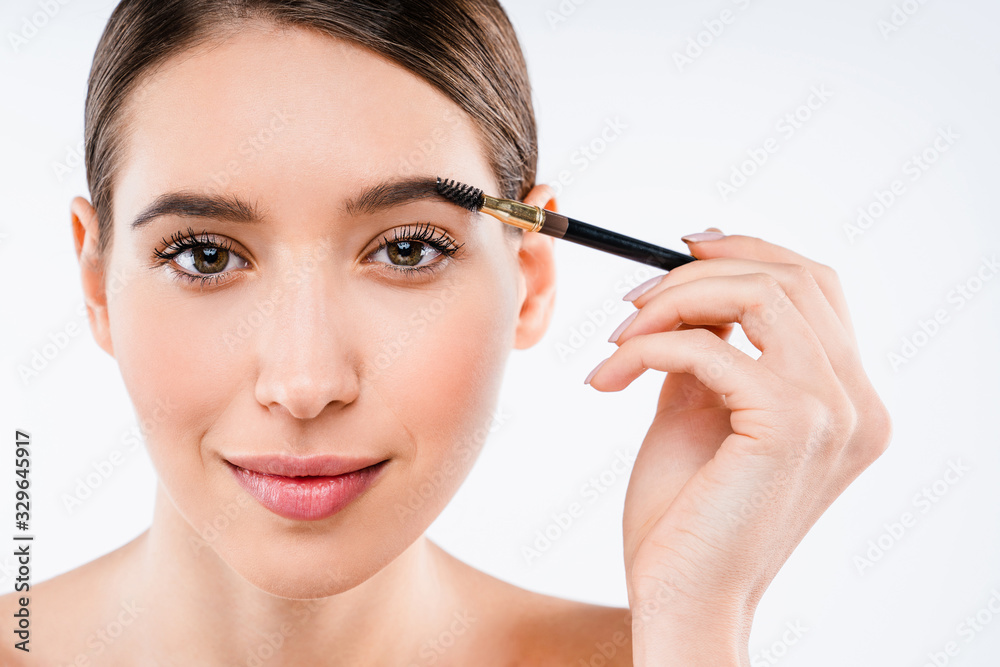 Close up of beautiful young woman applying makeup to her eyebrow while standing on isolated white background