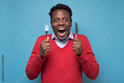 Young hungry african man holding a fork and a knife waiting for delicious meal. Isolated on blue background photo