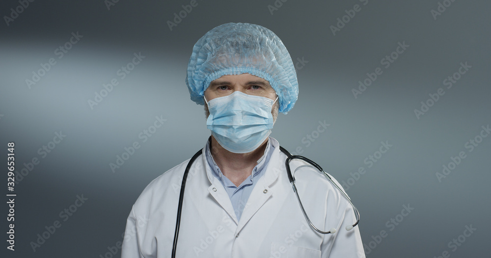 Portrait shot of the Caucasian handsome man doctor in blue hat taking off mask from his mouth and smiling cheerfully to the camera. Portrait shot of the Caucasian handsome man doctor in blue hat taking off mask from his mouth and smiling cheerfully to the camera.