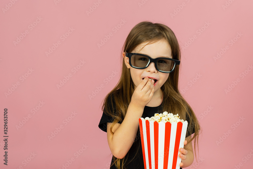 Fototapeta premium Cute little girl eats popcorn from striped packaging and watches a movie with 3D glasses. Cinema advertising concept, watching online movie