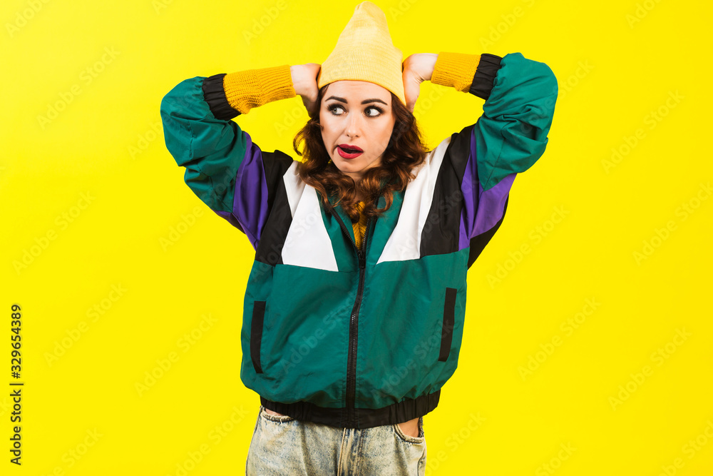 Fototapeta premium Modern beautiful girl in oversized sports jacket, hat and jeans, Billie Eilish style, back in the 90s, 2000s. Posing in the studio on a yellow background, strong funny emotions