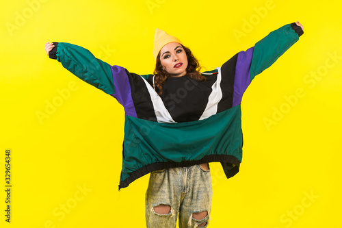 Modern beautiful girl in oversized sports jacket, hat and jeans, Billie Eilish style, back in the 90s, 2000s. Posing in the studio on a yellow background. photo