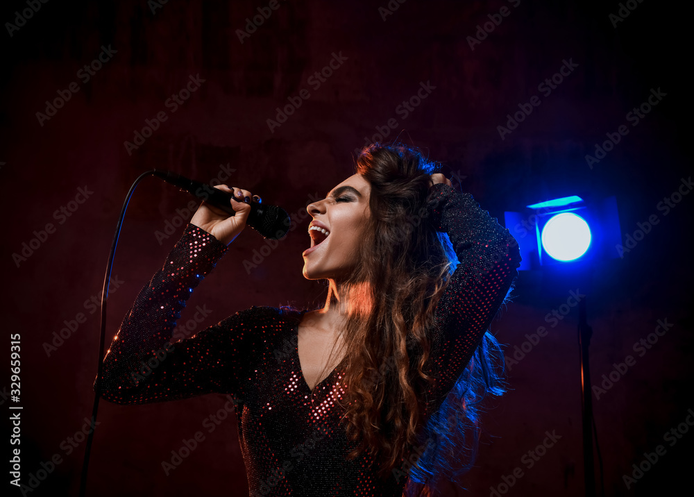 Beautiful singing girl curly afro hairstyle. Beauty woman singer sing with microphone karaoke song on stage in dark background Beautiful singing girl curly afro hairstyle. Beauty woman singer sing with microphone karaoke song on stage in dark background