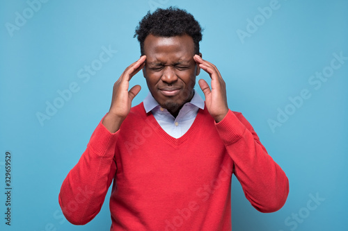 фотографія Serious afro american man in red sweater with headache in studio on blue isolate