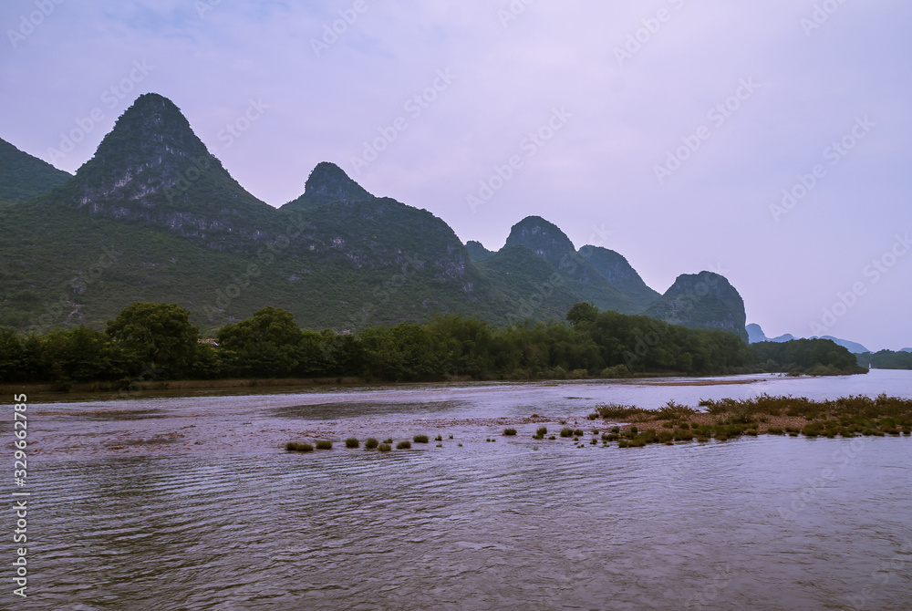 Guilin, China - May 10, 2010: Along Li River. Landscape of karst mountain under blueish cloudscape with green belt of trees on shoreline and dirt island up front.