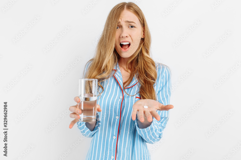 Portrait of a suffering young woman in pajamas holding a pill and a glass of water standing on a white background Portrait of a suffering young woman in pajamas holding a pill and a glass of water standing on a white background