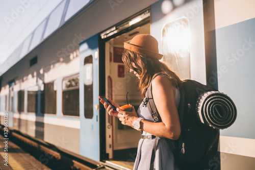 Railroad theme. Beautiful young woman with a backpack uses the phone while standing near the railroad train on the platform. Cheap travel photo
