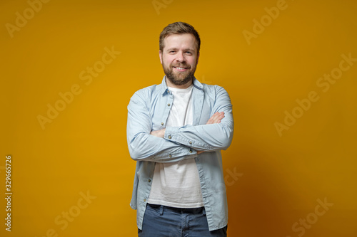 Confident bearded man stands with his arms crossed, cute smiling and looks at the camera with narrowed eyes.