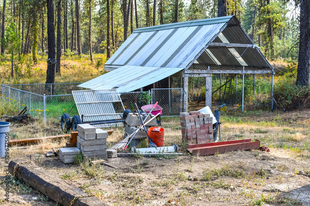 Tools for garden and construction materials sit on a dirt lot with a greenhouse behind in a rural setting.