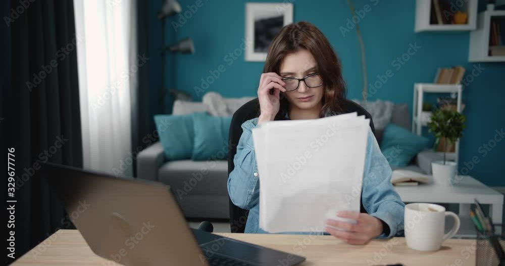 Beautiful young woman in eyeglasses and jeans shirt reading documents while sitting at table with personal laptop. Confident lady with curly hair working at home with modern interior. Beautiful young woman in eyeglasses and jeans shirt reading documents while sitting at table with personal laptop. Confident lady with curly hair working at home with modern interior.