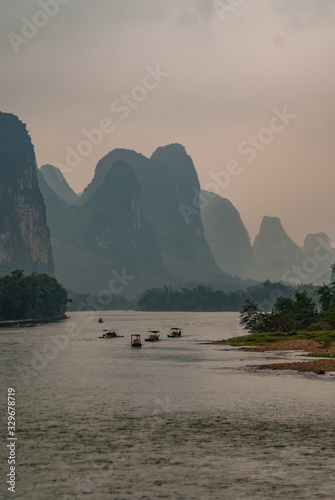 Guilin, China - May 10, 2010: Along Li River. Landscape with 5 small boats approaching on water with karst mountain range in back under brown cloudscape. Green trees on shoreline.