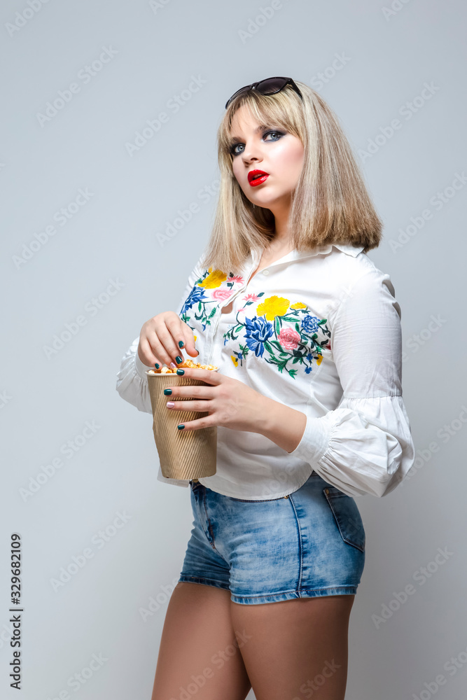 Portrait of Caucasian Female Blond Girl In Shorts Holding Popcorn Bucket and Posing Against White. Portrait of Caucasian Female Blond Girl In Shorts Holding Popcorn Bucket and Posing Against White.