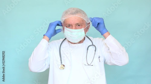 Elderly doctor putting on a mask, with white coat and blue gloves on light blue background. Old physician wearing protective equipment against Coronavirus Sars-CoV-2 spread and infectious diseases