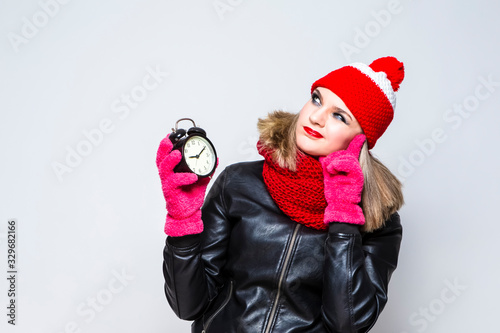 Time Concepts. Portrait of Exclaiming  Caucasian Blond Girl In Warm Hat and Scarf Posing with Round Clock With Thoughtful Expression Against White. photo