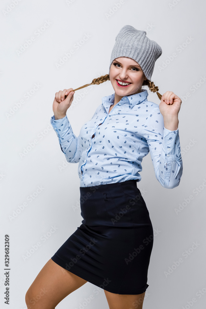 Youth Lifestyles. Portrait of Happy Caucasian Blond Girl in Warm Winter Hat and Blue Shirt Posing With Stretched Hair Tails on White Background. Youth Lifestyles. Portrait of Happy Caucasian Blond Girl in Warm Winter Hat and Blue Shirt Posing With Stretched Hair Tails on White Background.