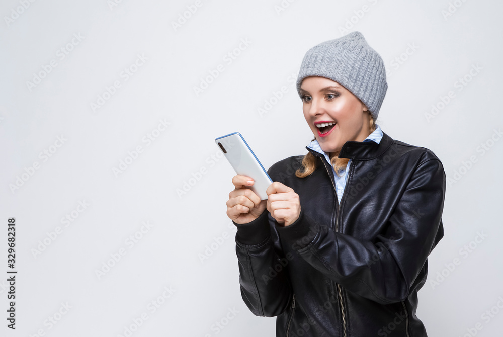 Youth Lifestyles.Excited Caucasian Blond Femalr in Warm Hat and Leather Jacket Posing With Cellphone on White Background Youth Lifestyles.Excited Caucasian Blond Femalr in Warm Hat and Leather Jacket Posing With Cellphone on White Background