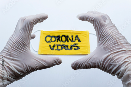 Closed up to White Esterilized gloves making an angry hand expression over yellow face mask with Black CORONAVIRUS inscription with white blackground. COVID-2019 photo