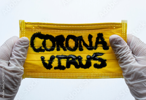 Closed up to Yellow Face Mask with CORONAVIRUS inscription and two hands with White Gloves or Esterilized globes holding them. COVID-2019 photo