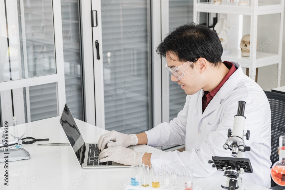 A male scientist with black hair wearing white coat and protective glassware sitting working on laptop with a microscope in white laboratory room with test tube and solutions.