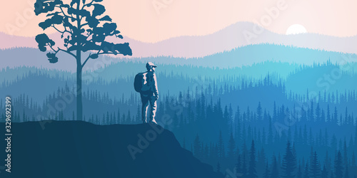 A tourist with a backpack on a rock meets dawn. Hills and mountains covered by forest, taiga. Morning haze, the light of sunrise.