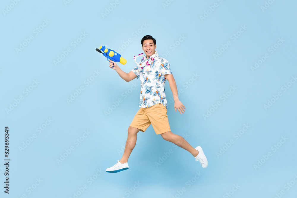 Handsome smiling young Asian man playing with water gun and jumping in studio blue background for Songkran festival in Thailand and southeast Asia Handsome smiling young Asian man playing with water gun and jumping in studio blue background for Songkran festival in Thailand and southeast Asia
