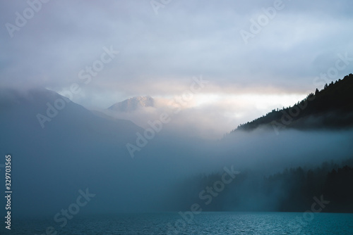 Beautiful mountain lake among huge mountains and forest in mist in golden hour. Sun shines through dense low clouds. Big rock glitters at sunrise. Alpine relaxing scenery with fog in pastel tones.