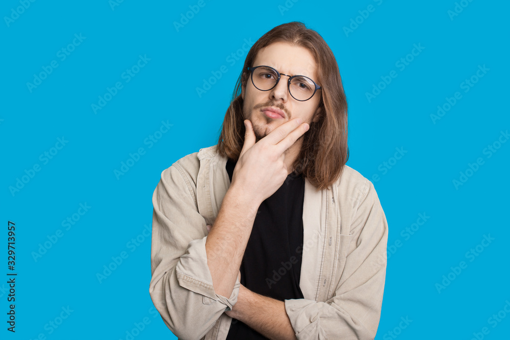 Thoughtful caucasian man with long hair and beard is looking through eyeglasses and touching his chin while posing on a blue wall Thoughtful caucasian man with long hair and beard is looking through eyeglasses and touching his chin while posing on a blue wall