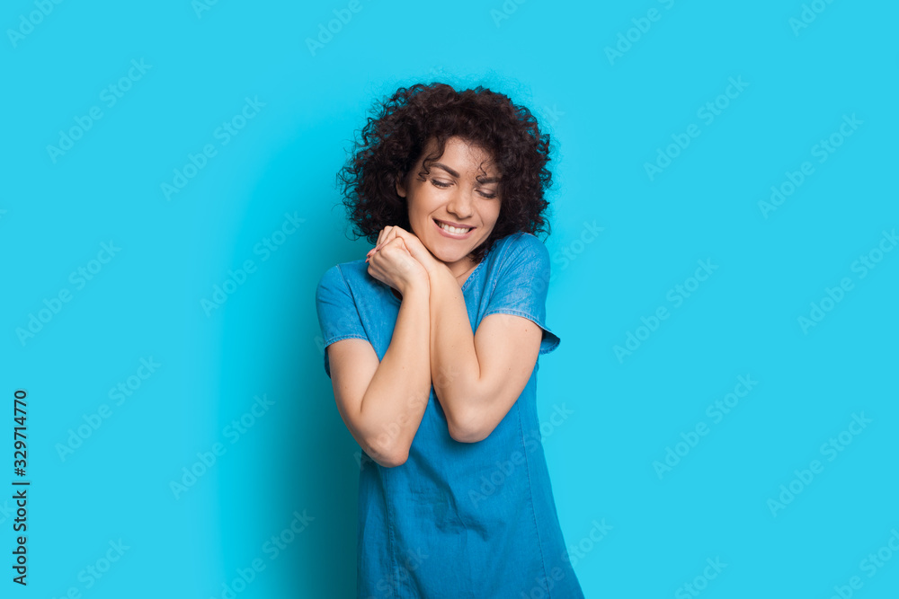 Curly haired caucasian lady feeling pleased on a blue background while posing in a dress Curly haired caucasian lady feeling pleased on a blue background while posing in a dress