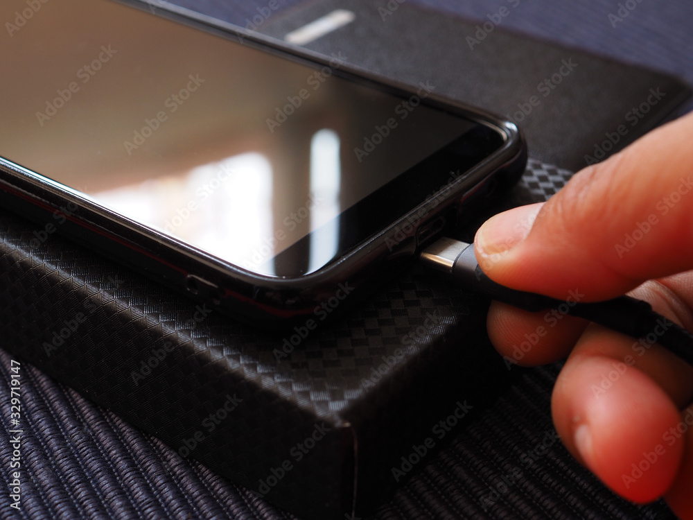 The hand is plugging the phone charger from the power bank on a black box over a blue background. Close up. The hand is plugging the phone charger from the power bank on a black box over a blue background. Close up.
