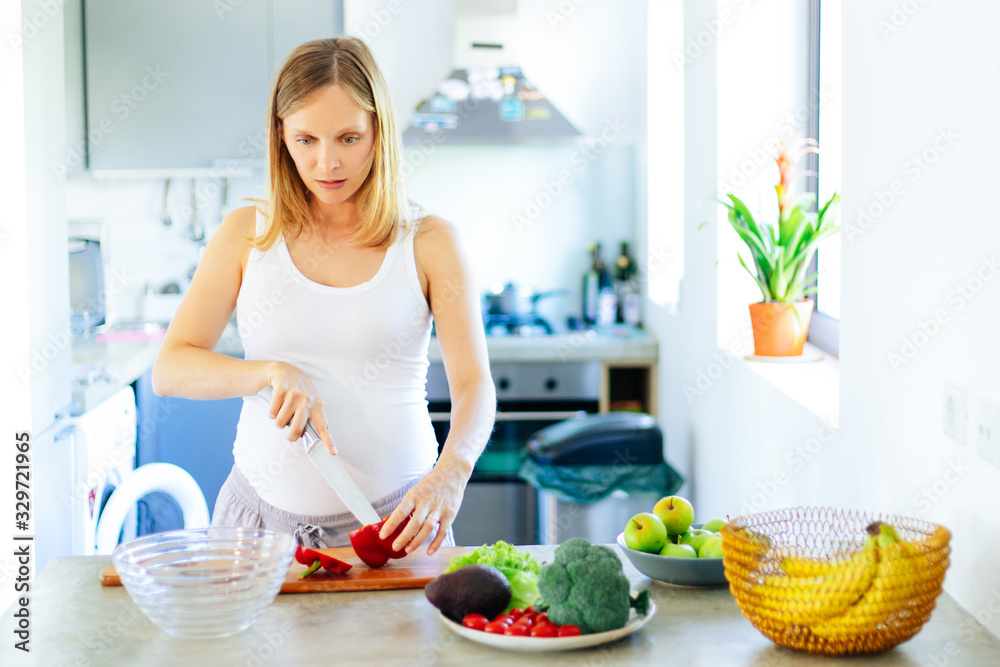 Pensive serious pregnant woman cooking salad in kitchen. Young expectant mother standing at table with bowls of vegetables, cutting pepper. Organic food concept Pensive serious pregnant woman cooking salad in kitchen. Young expectant mother standing at table with bowls of vegetables, cutting pepper. Organic food concept