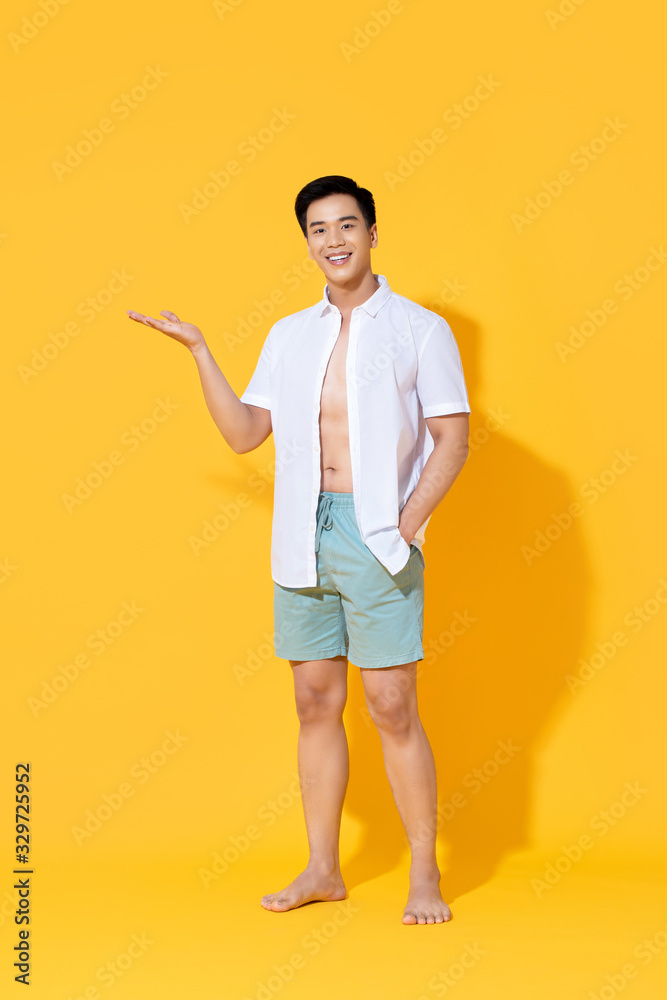 Young smiling handsome Asian man in beach attire doing open palm gesture showing blank space in summer yellow isolated studio background Young smiling handsome Asian man in beach attire doing open palm gesture showing blank space in summer yellow isolated studio background