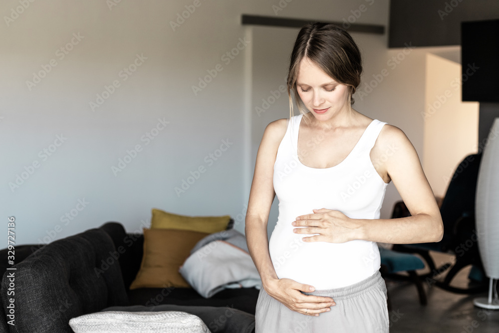 Peaceful expectant mother standing in living room. Pregnant young woman holding baby bump, smiling, looking away. Pregnant woman at home concept