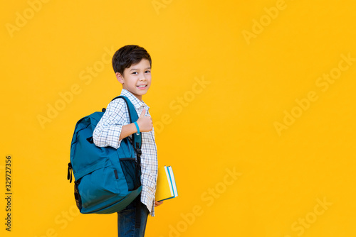 Smiling handsome mixed race schoolboy with books and backpack giving thumbs up isolated on yellow background with copy space photo