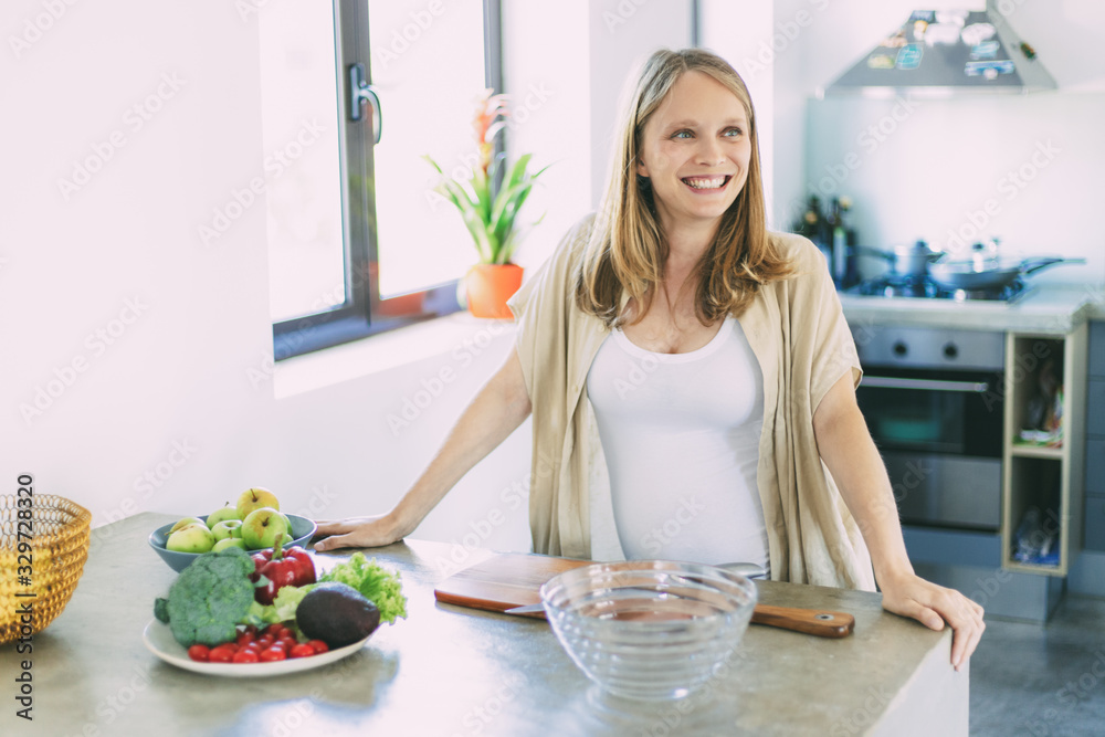 Joyful expectant mother cooking salad in kitchen. Young pregnant woman standing at table with bowls of vegetables, smiling, looking away. Pregnancy and nutrition concept