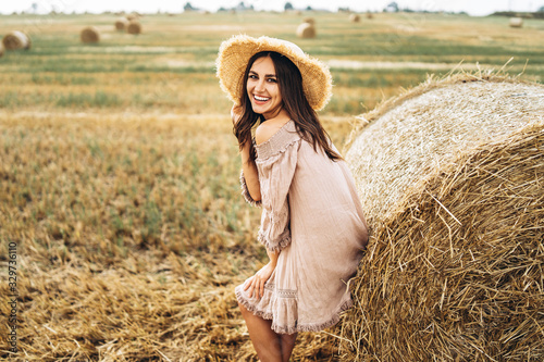 Beautiful young girl with long hair in sunnglasses and straw hat posing on a wheat field near hay bales. Happy brunette in summer dress © bedya