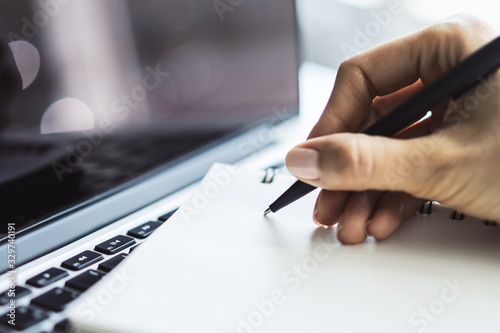 Photos Woman writes with a pen in notebook on laptop keyboard in a sunny office, business and education concept