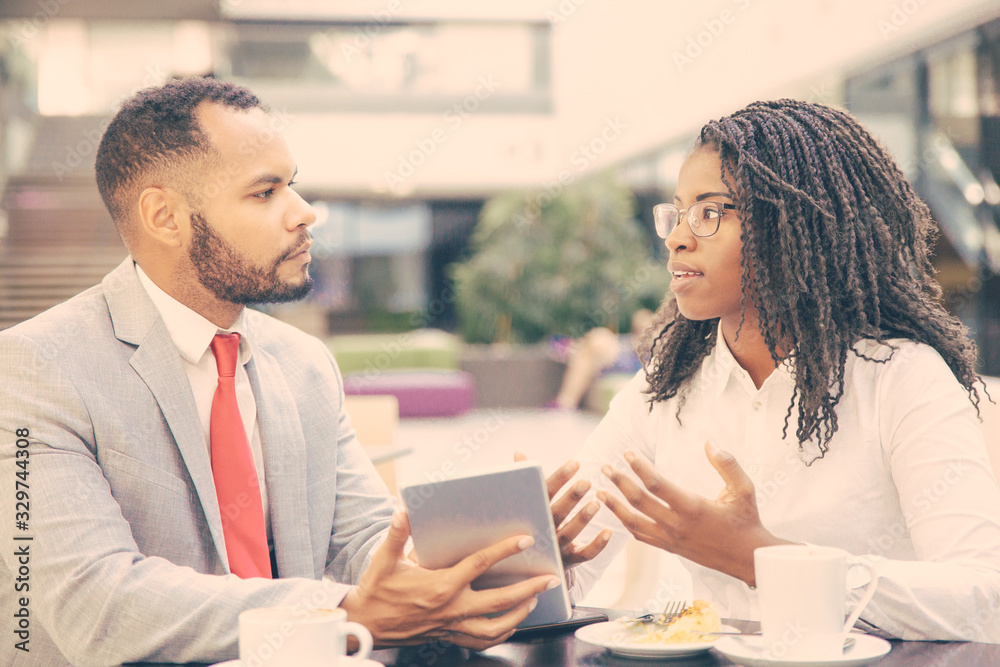 Excited business colleagues discussing tablet app. Diverse business man and woman sitting in cafe, using tablet together and talking. Digital communication concept