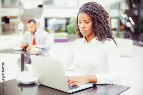 Confident young businesswoman using computer in co-working space. African American business woman looking at laptop screen, man using tablet in background. Young businesswoman concept