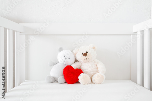 Two smiling white teddy bears sitting in baby bed. Red fluffy heart in middle. Front view. Closeup.