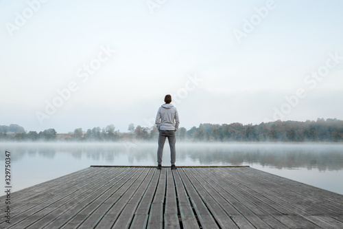 Young man standing alone on wooden footbridge and staring at lake. Thinking about life. Mist over water. Foggy air. Early chilly morning. Peaceful atmosphere in nature. Enjoying fresh air. Back view.