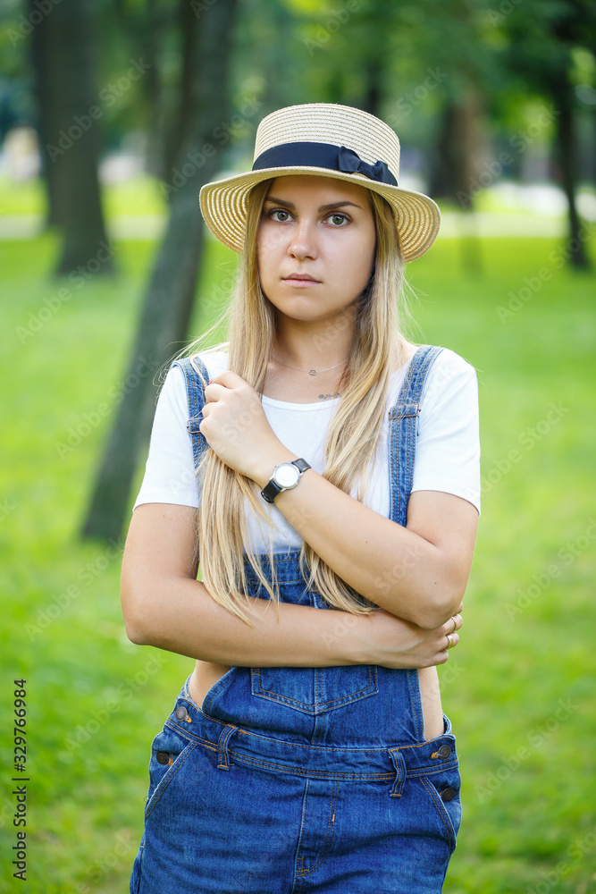 young beautiful girl in denim overalls and a light hat walking in the park