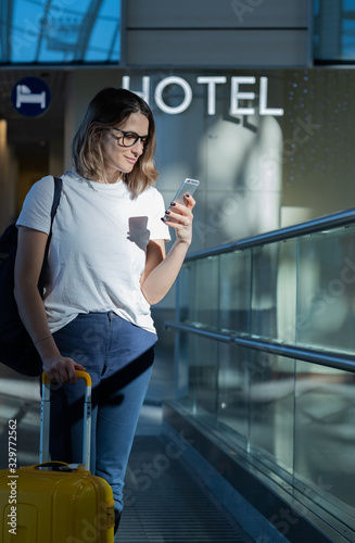 A beautiful business woman stands at the airport with a yellow suitcase and writes a message