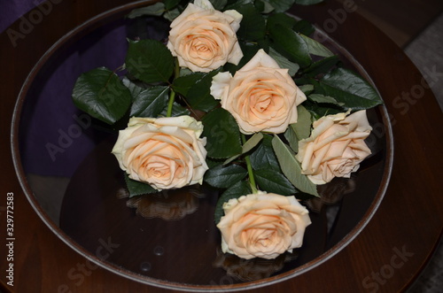  Against the background of purple textile, on a round glass table is a bouquet of beautiful fresh fragrant orange roses with green leaves.