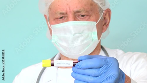 Worried elderly doctor analysing positive test tube of Coronavirus, Covid-19. Old physician looking infected blood examination of Sars-CoV-2 with white coat and blue gloves on light blue background