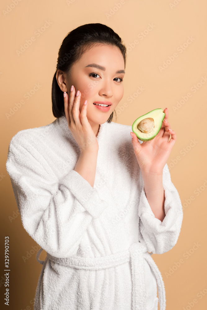 beautiful asian woman in bathrobe with avocado half touching face on beige background beautiful asian woman in bathrobe with avocado half touching face on beige background