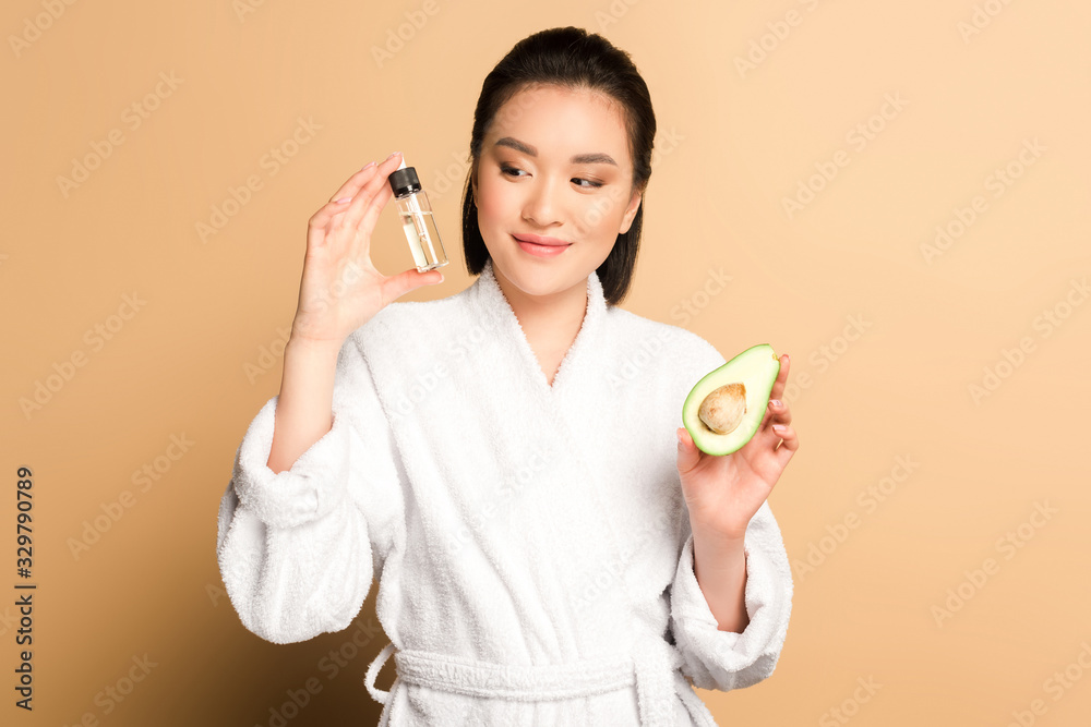 happy beautiful asian woman in bathrobe with avocado half and oil in bottle on beige background happy beautiful asian woman in bathrobe with avocado half and oil in bottle on beige background