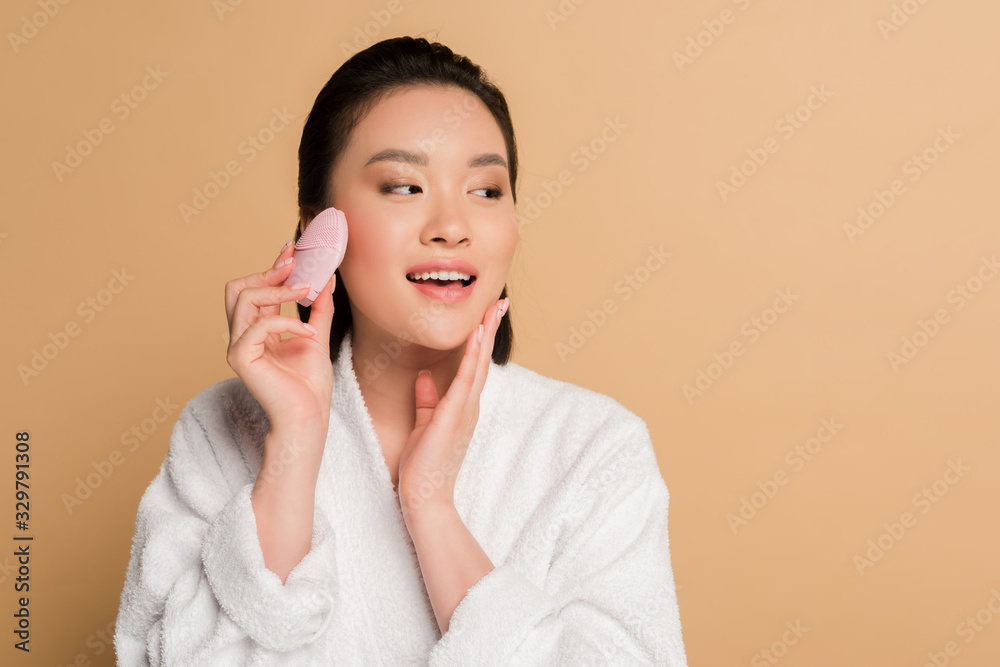 beautiful asian woman in bathrobe using facial cleansing brush on beige background