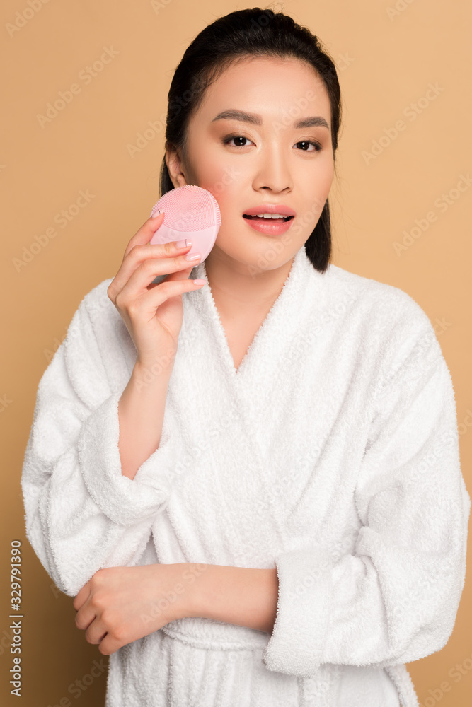 beautiful asian woman in bathrobe using facial cleansing brush on beige background beautiful asian woman in bathrobe using facial cleansing brush on beige background